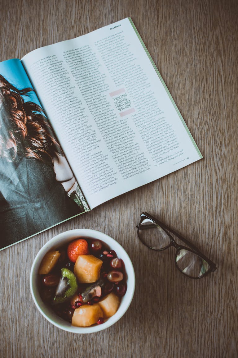 A fresh fruit bowl, open magazine, and glasses on a wooden table, perfect for lifestyle and food content.